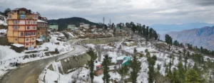 Snow-covered landscape in Shimla with hillside houses, winding mountain road, and pine trees during winter snowfall in Himachal Pradesh