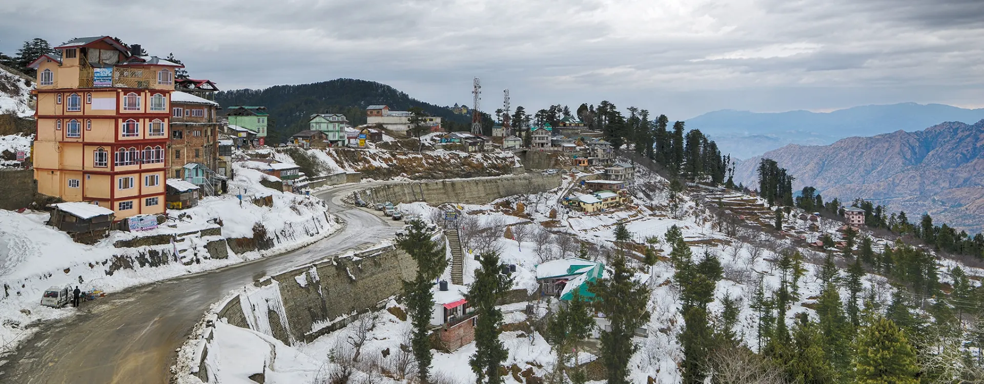 Snow-covered landscape in Shimla with hillside houses, winding mountain road, and pine trees during winter snowfall in Himachal Pradesh